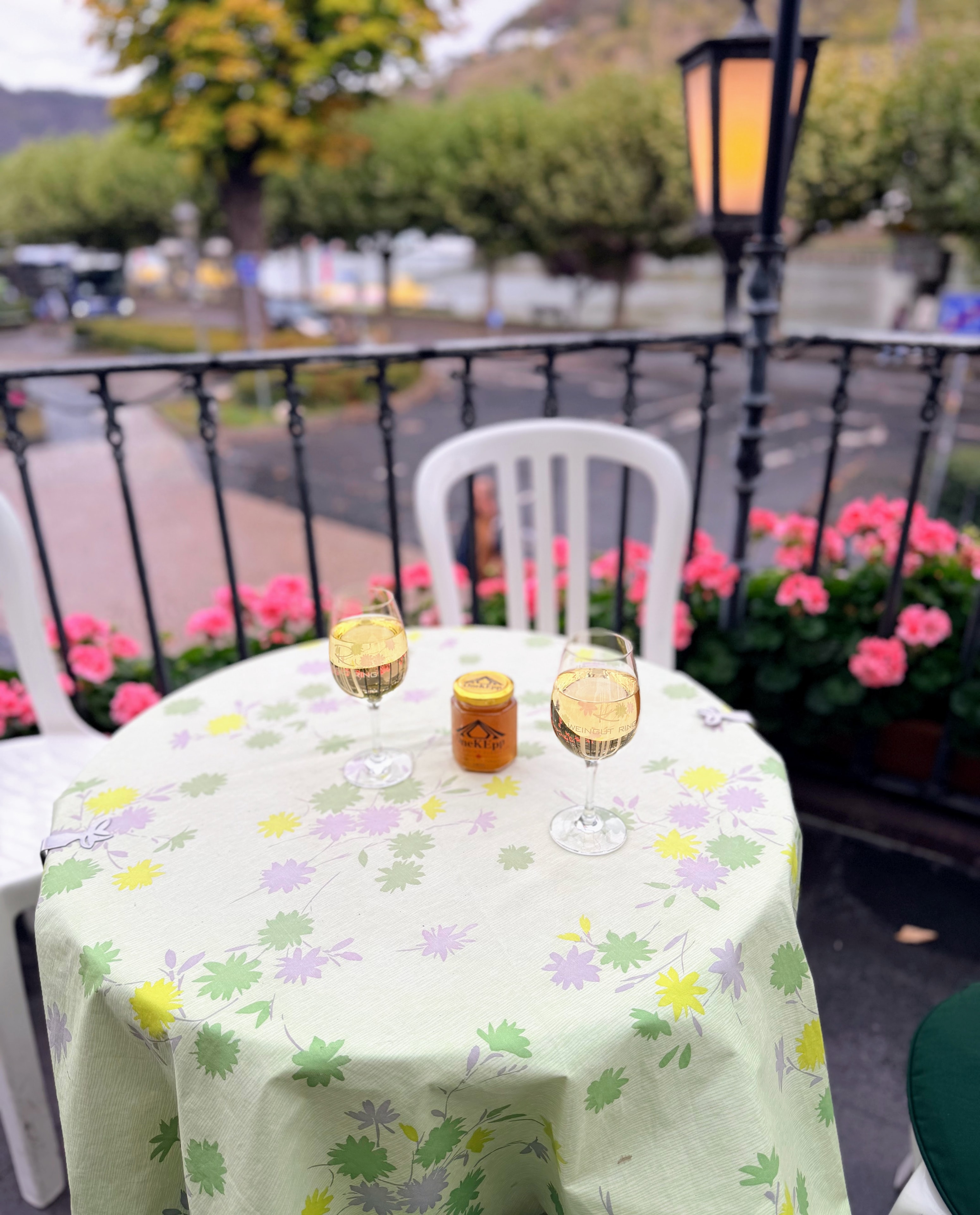 OneKEpp honey jar between two glasses of Riesling on a floral tablecloth balcony table with pink geraniums