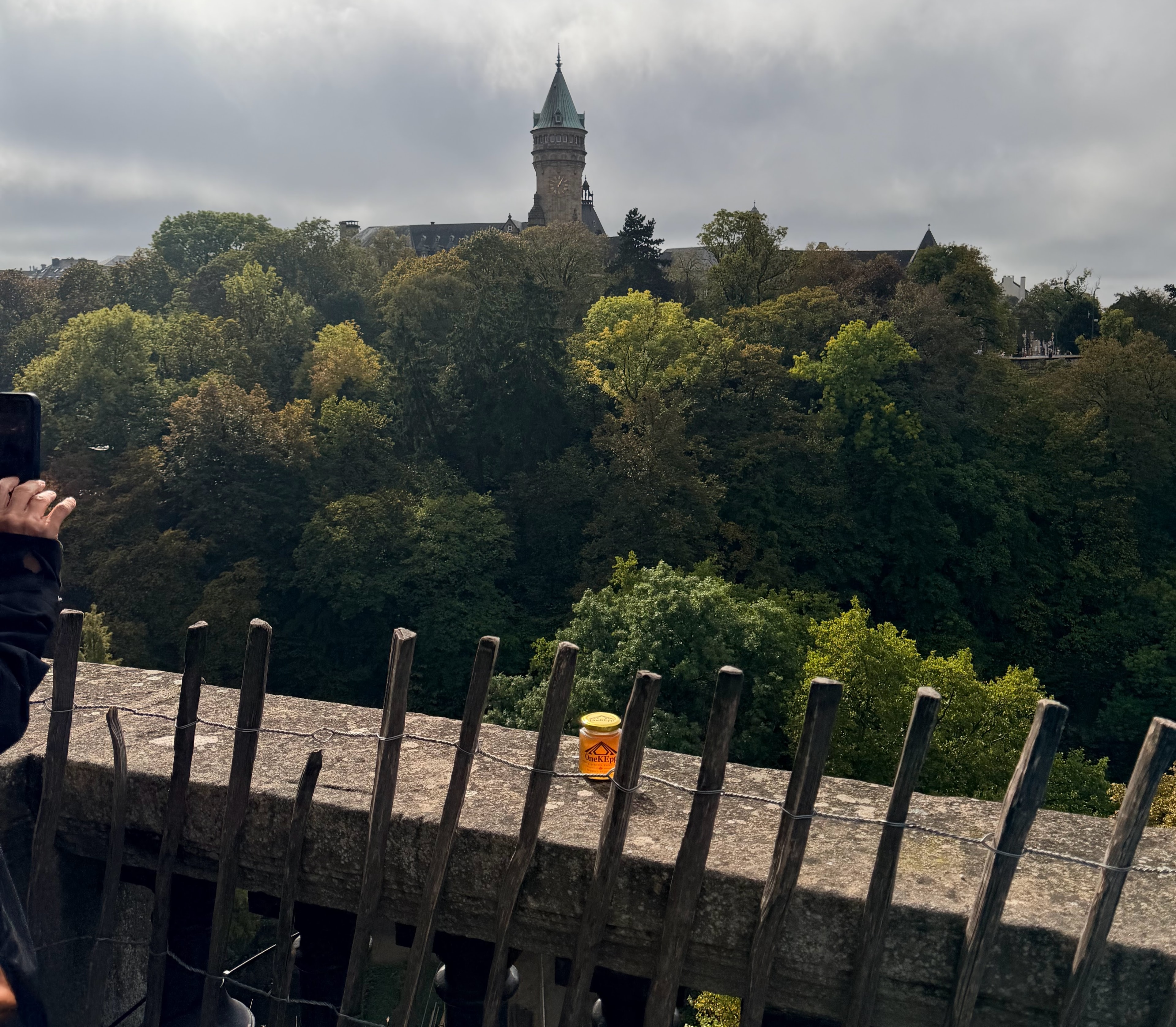 OneKEpp honey jar on a stone wall with the Luxembourg State Savings Bank tower rising above the forest